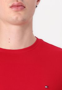 Červené tričko s kulatým výstřihem vyrobené z bavlny, s texturovaným povrchem a malým vyšitým logem v modré, bílé a červené barvě na dolní levé straně.