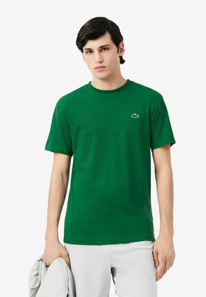 Jonge man die een groen T-shirt met krokodillenlogo en lichte korte broek draagt, met een lichtgrijs jasje in zijn linkerhand.