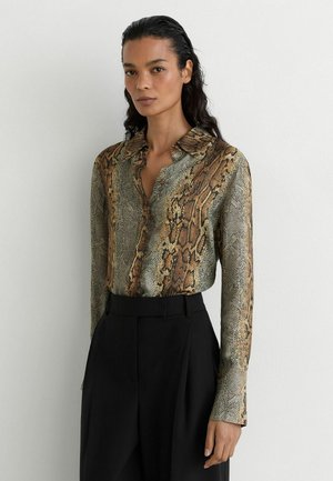 Vrouw met donker golvend haar draagt een bruine en groene blouse met slangenprint en lange mouwen, ingestopt in een zwarte broek met hoge taille, tegen een effen achtergrond.