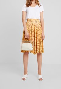 Keltainen kukallinen laskostettu hame, valkoinen V-kaula-aukkoinen T-paita, läpinäkyvä laukku, jossa on kerma pohja ja puinen kahva, valkoiset ristisäädettävät sandaalit.