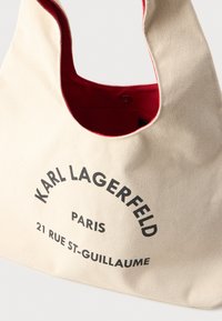 Bežová plátenná taška s červenou podšívkou a čiernym textom "Karl Lagerfeld Paris 21 Rue St-Guillaume" na prednej strane.