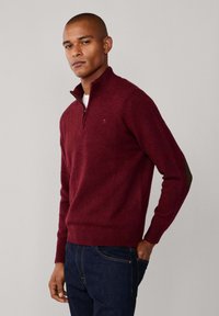 Pullover lavorato a maglia color bordò con colletto alto, caratterizzato da una chiusura a mezza zip e polsini a costine. Abbinato a jeans denim blu scuro.