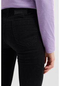 Černé džíny z denimu s zadní kapsou a viditelným švem, nošeny s dlouhým rukávem v světle fialové barvě, ukazující část ruky.