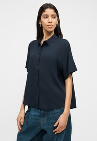 Γυναίκα με φαρδιά σκούρα μπλε κοντομάνικη πουκάμισα με κουμπιά και φαρδιά μπλε τζιν παντελόνια, στέκεται με το ένα χέρι στην τσέπη μπροστά σε λευκό φόντο.