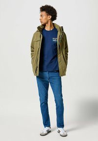 Olivenfarvet jakke med hætte, marineblåt skjorte, blå jeans og hvide sneakers med grå og sorte accenter. Enkel design, funktionelle lommer.