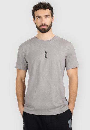 Mężczyzna z kręconymi włosami i brodą, ubrany w jasnoszary, wzorzysty t-shirt z małym czarnym logo i czarne spodnie, ręce w kieszeniach, neutralne tło.