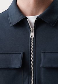 Giacca blu navy con zip realizzata in un tessuto morbido e liscio. Presenta un collo e due tasche sul petto con pattina. Dettagli in zip argentata.