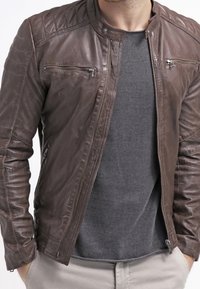 Uomo con una giacca di pelle marrone sopra un maglione grigio, con le mani nelle tasche dei pantaloni beige chiaro, su uno sfondo bianco.