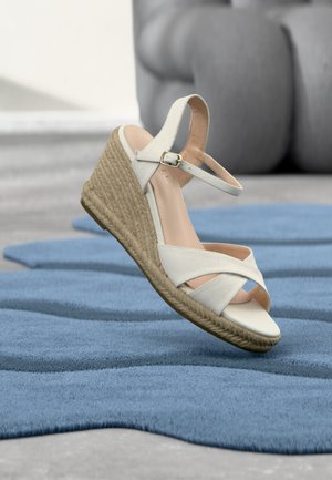 Weißer Keilsandalette mit Knöchelriemen und geflochtener Sohle, die über einem blau strukturierten Teppich schwebt, mit einem grauen Sofa im unscharfen Hintergrund.