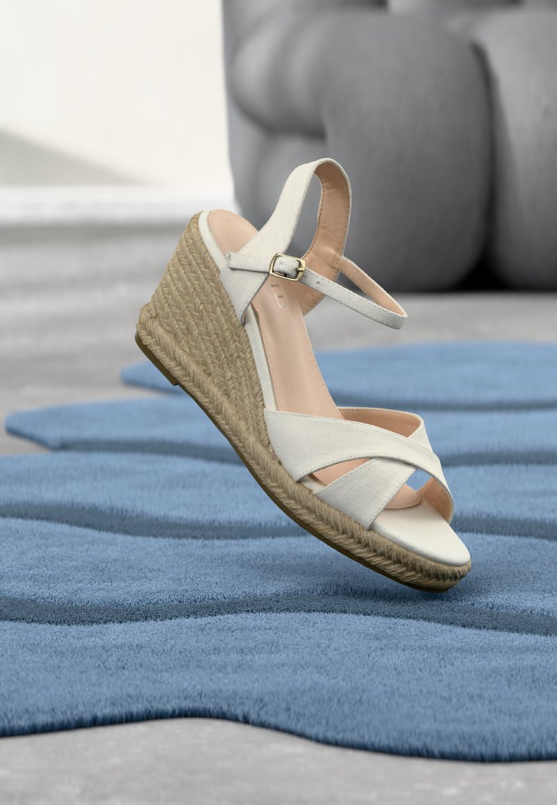 Sandale compensée blanche avec bride à la cheville et semelle tressée suspendue au-dessus d'un tapis texturé bleu, avec un canapé gris en arrière-plan flou.