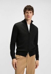 Svart zip-up cardigan med hög krage, gjord av texturerat tyg. Har ribbade ärmslut och rak fåll, bärs över en vit t-shirt.