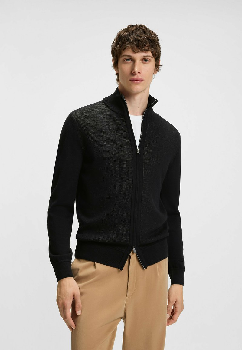 Svart zip-up cardigan med hög krage, gjord av texturerat tyg. Har ribbade ärmslut och rak fåll, bärs över en vit t-shirt.