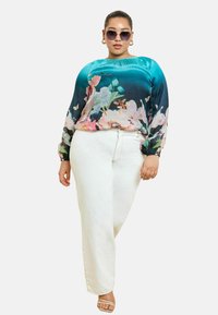 Blusa floreale con colori turchesi e rosa, vestibilità ampia, scollatura elasticizzata e maniche lunghe, abbinata a jeans bianchi a palazzo e sandali con cinturini.