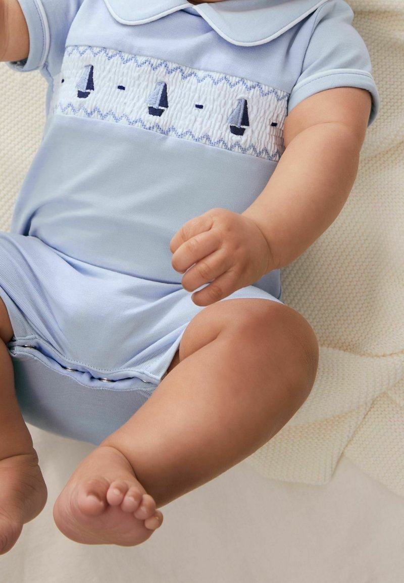 Barboteuse en coton bleu clair avec un motif de voilier blanc brodé sur la poitrine. Manches courtes et fermeture à boutons pression au bas.