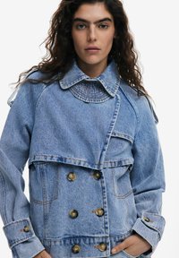 Jachetă din denim de culoare bleu deschis, cu design dublu-breasted, guler mare, cusături vizibile și accente cu butoane pe mâneci și față.