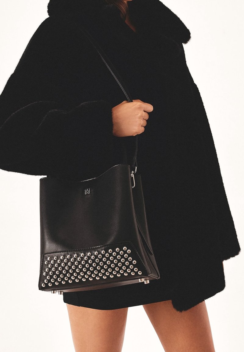 Personne portant un manteau noir et une jupe courte tenant un sac à main en cuir noir avec des clous argentés sur le bord inférieur.