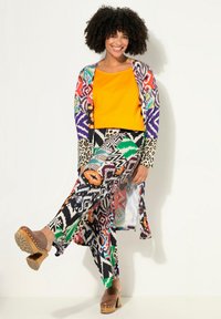 Kleurige outfit met een feloranje top, een patroonrijke lange cardigan en bijpassende broeken met verschillende geometrische en dierenprints. Bruine klompen.