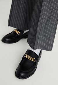Black leather loafers with a gold chain detail across the front, paired with gray pinstripe trousers and light patterned socks.