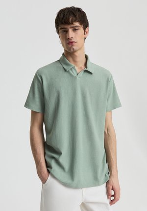 Giovane uomo con polo verde chiaro a maniche corte e pantaloni bianchi, in piedi davanti a uno sfondo chiaro e uniforme.