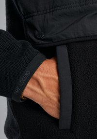 Chaqueta negra con una manga de tela texturizada y un cuerpo de forro polar suave, con un logo en el puño y un bolsillo en la parte delantera.