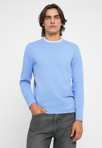 Suéter azul claro de manga longa com decote redondo. Textura suave, forma ajustada, combinado com jeans cinza escuro, mãos nos bolsos.