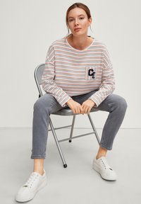 Camicia a manica lunga a righe con colori grigio, arancione e bianco; presenta una tasca con dettaglio ricamato; abbinata a jeans grigi e sneakers bianche.