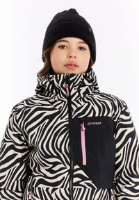 Takki, jossa on seeprahiihtokuosi ja korkea kaulus, mustavalkoista kangasta, vaaleanpunaisia vetoketjujen yksityiskohtia sekä musta tasku, yhdistettynä neulottuun mustaan pipoon.