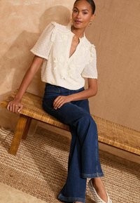 White textured blouse with floral embroidery, puff sleeves, and a v-neck. Paired with dark blue flared jeans and white woven slip-on shoes.