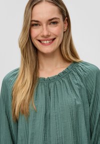 Lichtgroene geplooide blouse met een zachte rimpelnek en lange mouwen, gemaakt van een gestructureerde stof.