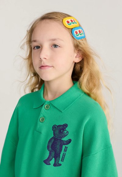 Jeune fille aux longs cheveux blonds porte un polo vert avec un imprimé ours en peluche et des barrettes à fleurs colorées sur un fond clair.