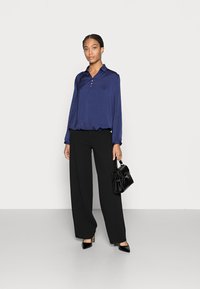 Blusa leve de cor azul marinho com gola e quatro botões, combinada com calças largas pretas de cintura alta e uma pequena bolsa preta.