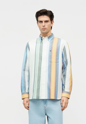 Homme portant une chemise à rayures verticales multicolores avec une poche poitrine, associée à un jean bleu clair, debout devant un fond uni.