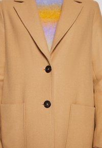 Beżowa wełniana kurtka z ząbkowanymi klapami, trzema czarnymi guzikami i dwiema przednimi kieszeniami. Pod nią widoczny jest wielokolorowy sweter.