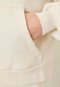 Hoodie in pile di colore crema con superficie testurizzata, dotato di tasca frontale e polsini a costine ai polsi e all'orlo.