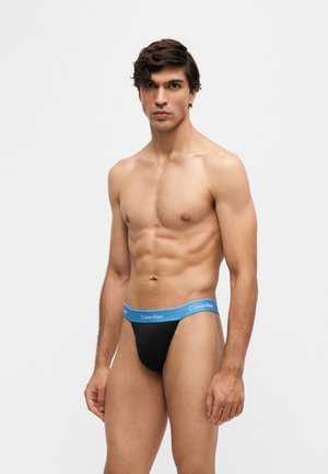 Jonge man met kort donker haar, gekleed in zwarte Calvin Klein slips met een blauwe tailleband, staand tegen een effen lichte achtergrond.