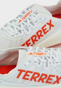Białe buty sportowe z czerwonymi akcentami, wykonane z siateczkowego materiału, z charakterystycznym systemem sznurowania i dużym logo "TERREX" po boku.