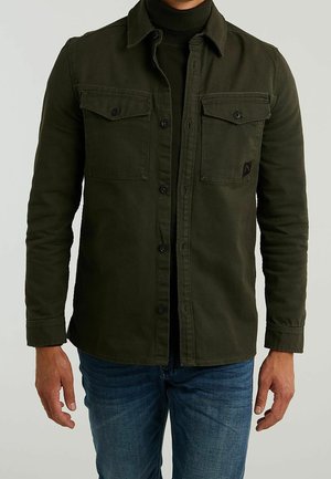 Homme portant une surchemise kaki à boutons sur un col roulé noir et un jean bleu, debout les bras détendus le long du corps.