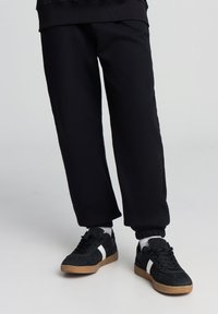 Pantaloni della tuta neri con vestibilità comoda e polsini elastici, abbinati a sneakers nere con una striscia laterale bianca e suola in gomma.