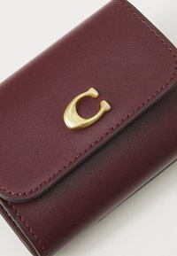 Portefeuille en cuir bordeaux avec un logo "C" doré, présentant des bords cousus et un design élégant et compact.