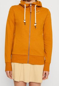 Felpa con zip arancione con cappuccio, dotata di lacci bianchi, tasche laterali e polsini a costine. Indossata sopra una gonna leggera beige.