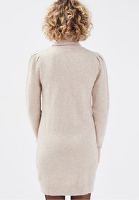 Beiges Strickkleid mit langen Ärmeln, hohem Kragen und dezenten, leicht gepufften Schultern; figurbetonte Silhouette und gerippter Saum.