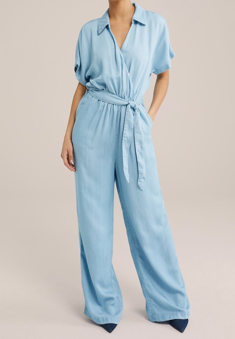 Ljusblå långärmad jumpsuit med V-ringning, knytlinning och vida byxor. Slät textur och avslappnad design.