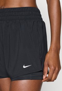 Sorte atletikshorts med en bred elastisk talje. Har et hvidt Nike-logo på nederste venstre side og en glat, letvægtsstoftekstur.
