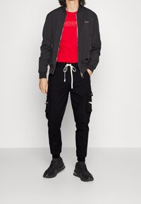 Svart bomberjacka med ribbade muddar, röd grafisk t-shirt, svarta cargobyxor med fickor och svarta sneakers med texturerad sula.