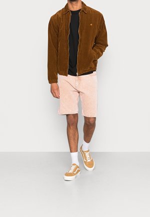 Ruskea corduroy-takki, musta t-paita, vaaleanpunaiset shortsit, valkoiset sukat, ruskeat lenkkarit. Sileä pinta; rento istuvuus; vetoketjulla suljettava takki.