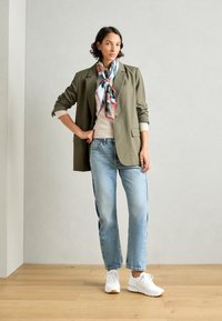 Blazer attillato verde oliva, maglione beige, jeans a gamba dritta blu chiaro e sneakers bianche con suola in gomma. Sciarpa di seta a fantasia colorata.