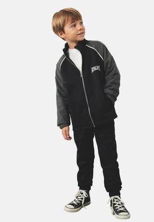 Chaqueta negra con cremallera y mangas grises, logo blanco, combinada con pantalones negros y zapatillas negras. Diseño simple, adecuado para uso casual.
