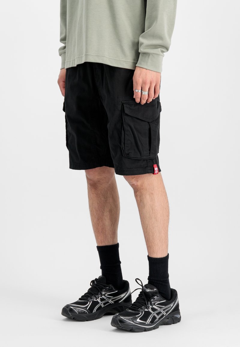 Personne portant un short cargo noir, des chaussures de sport noires, des chaussettes noires et une chemise à manches longues vert clair, debout devant un fond uni.