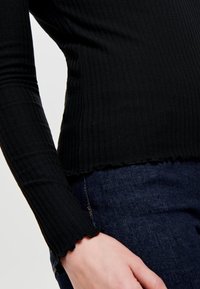 Haut noir côtelé à manches longues avec un ourlet festonné, porté avec un jean en denim foncé. Le tissu a une texture douce et une coupe ajustée.