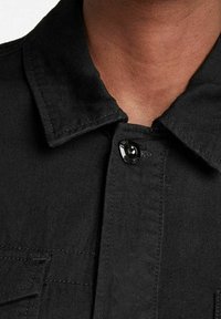 Närbild av en person som bär en svart skjorta med krage, knäppt upptill, med synlig sömnad och tygtextur.